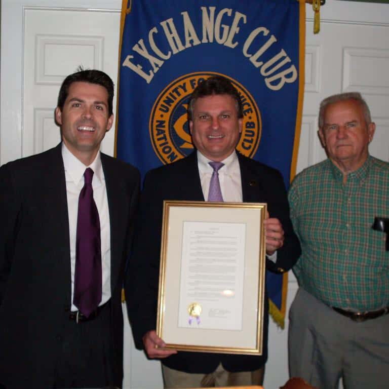 A smiling man in a jacket and tie holding a framed certificate of recognition with two gentlemen standing on either side of him