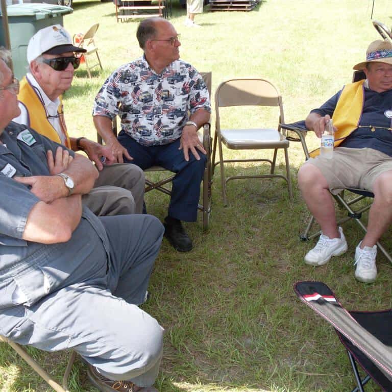 Four older men sitting on folding chairs under a tent