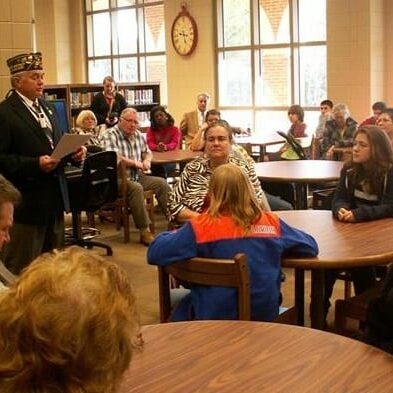 A gentleman speaker reading information to a group of people seated at round tables