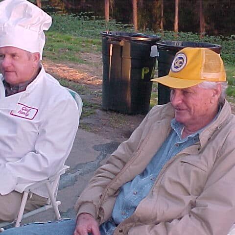 Two men outside sitting in folding chairs, one in a white chef's uniform and the other sporting an Exchange Club cap