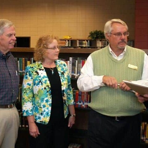 A woman and three men standing in a library listening to a man in a blazer reading from a clipboard