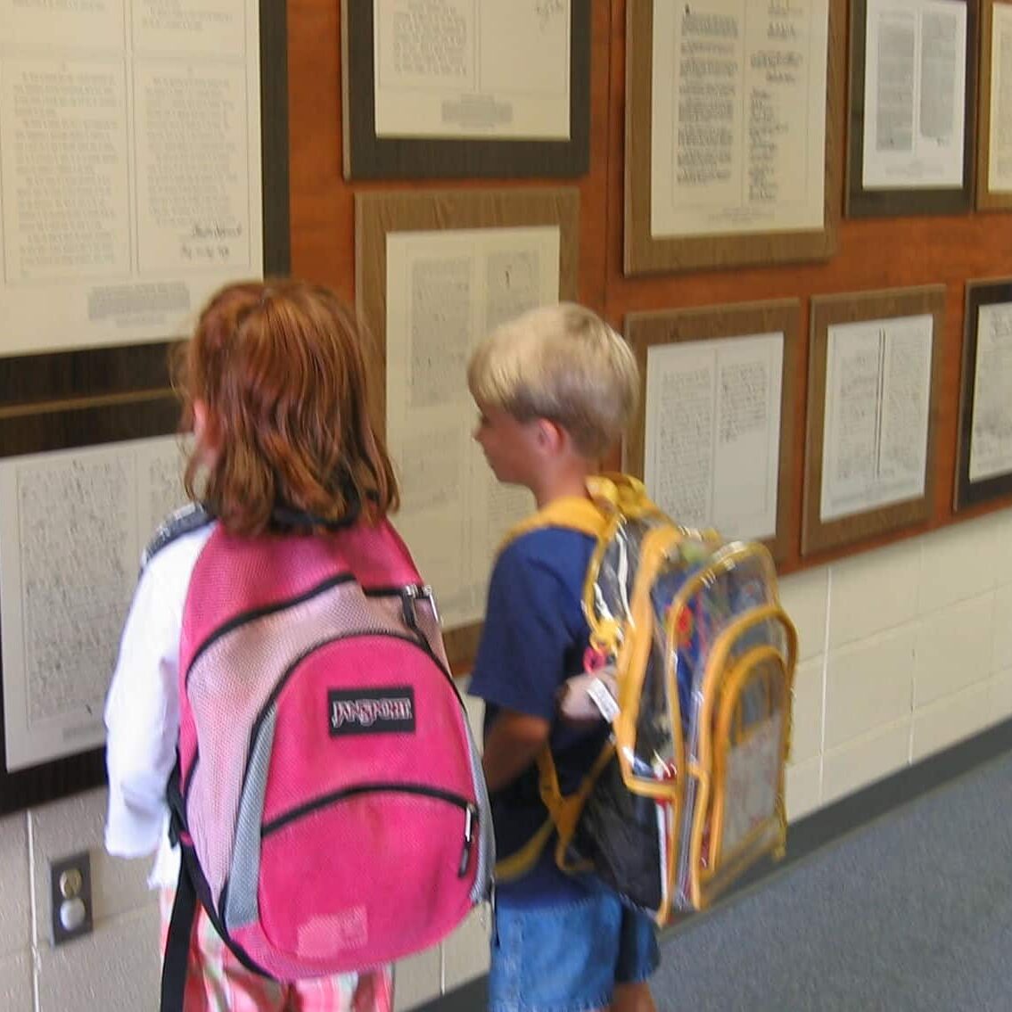 A girl and boy wearing school backpack while looking at a wall of framed documents