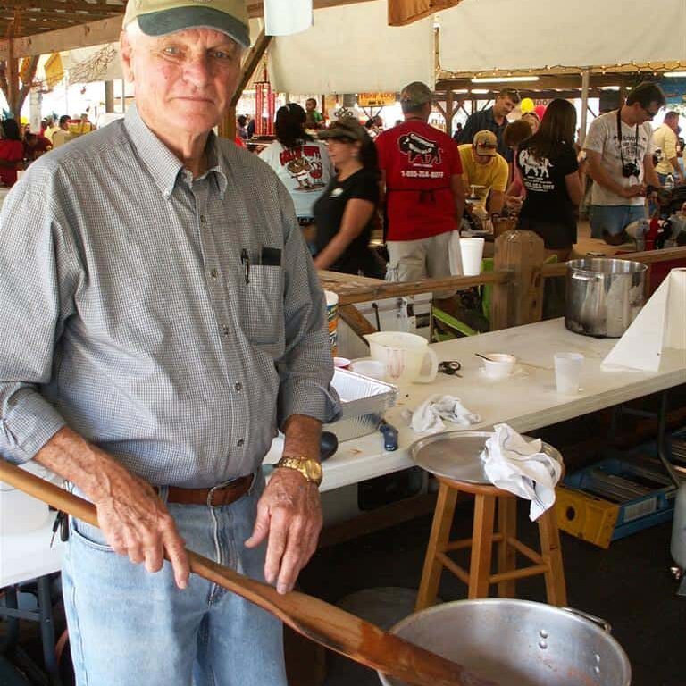 An older gentleman stirring a large aluminum pot with a big wooden paddle