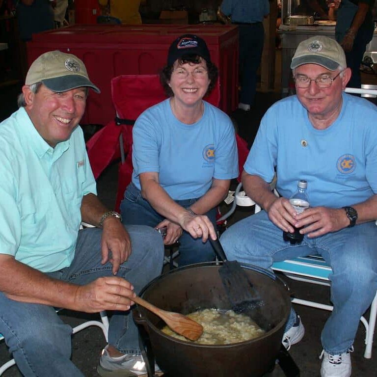 A woman and two men in Exchange Club shirts sitting in folding chairs while taking turns stirring a large cast iron pot