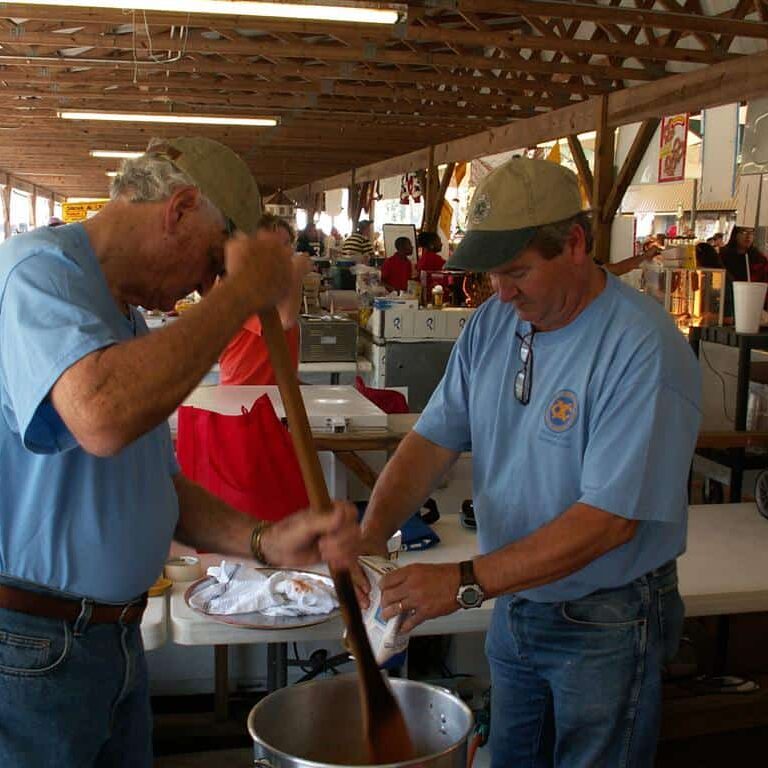 One man stirring a large aluminum pot with a long wooden paddle while the other adds ingredients