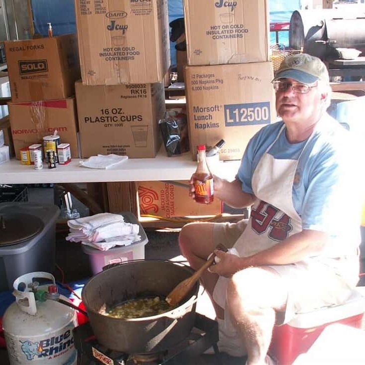 Man holding a bottle of hot sauce as he stirs a large cast iron pot on a propane cooker