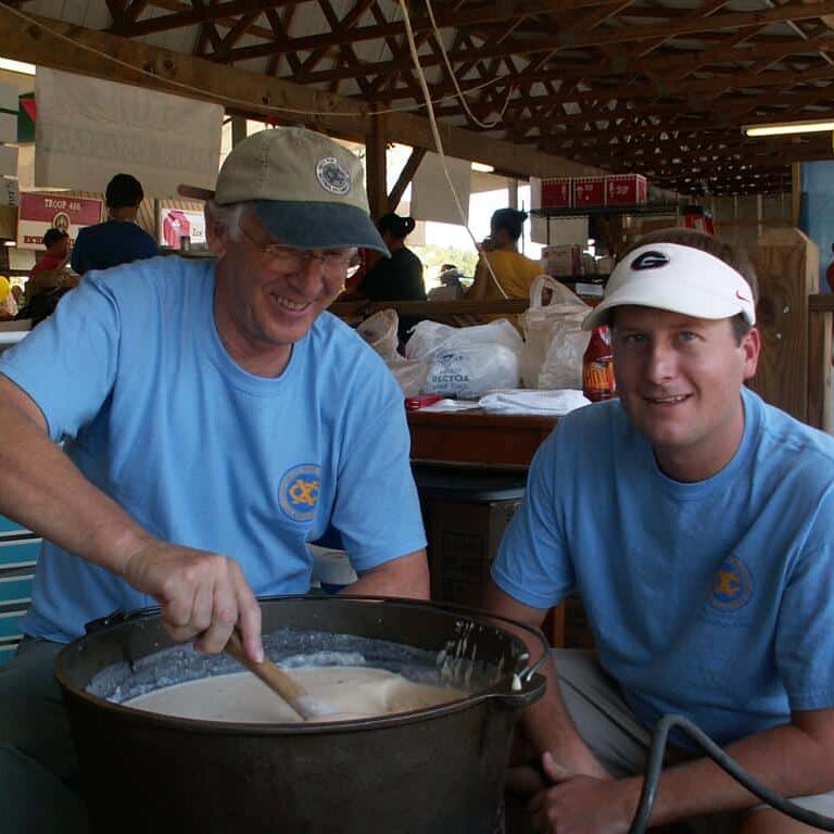 Two men wearing blue Exchange Club T-shirts seated while one stirs a large cast iron pot with a wooden spoon