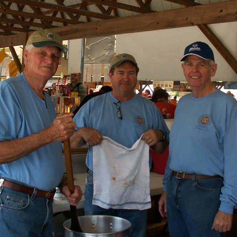 Three men in Exchange Club T-shirts tending to a large aluminum pot of food