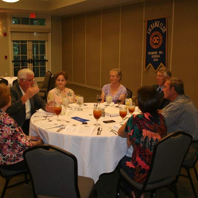 Seven men and women seated at a round banquet table talking