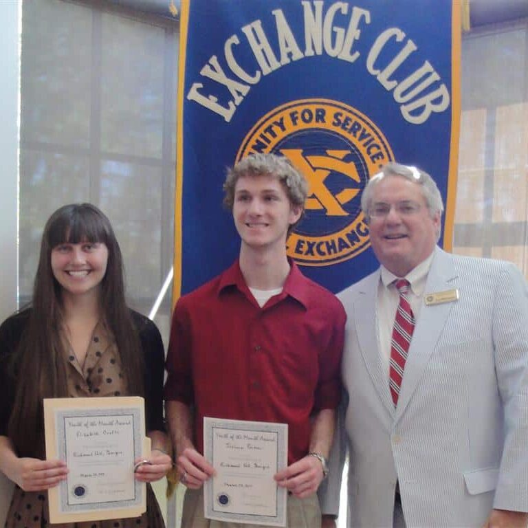 A young adult female and male student holding certificates presented by an Exchange Club member wearing a red striped tie and seersucker suit