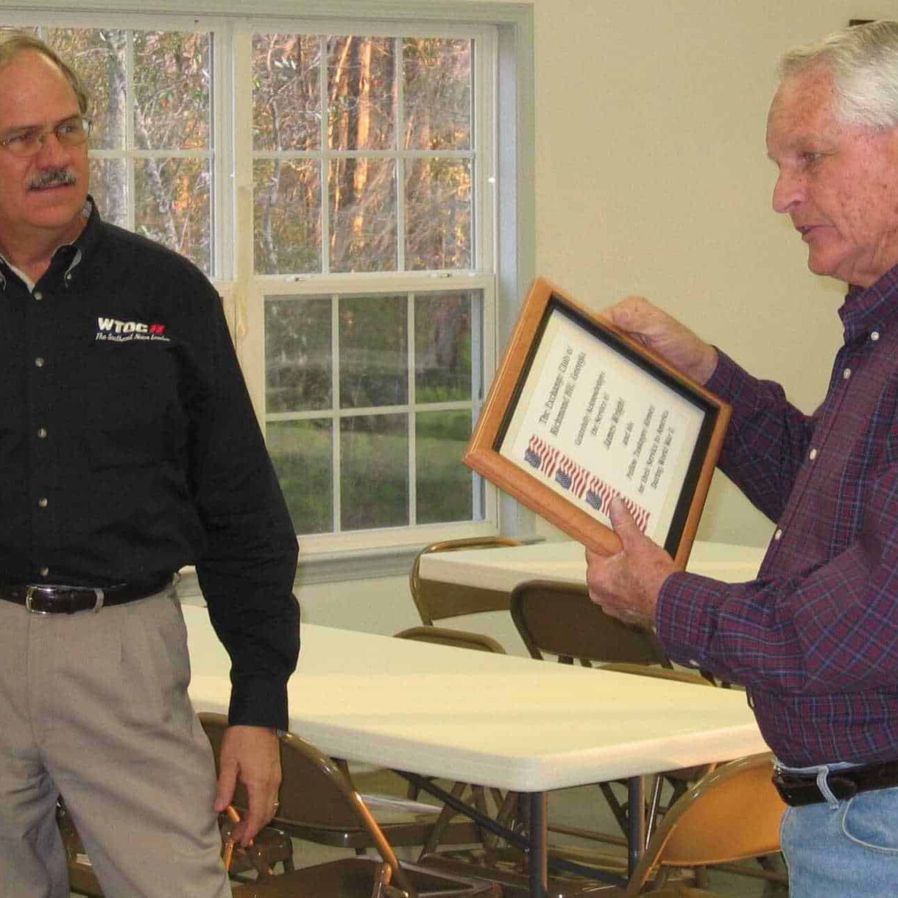 A man presents an older veteran with a framed Exchange Club acknowledgment in recognition of his military service