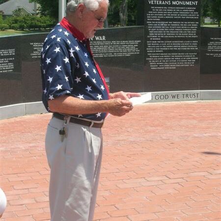 A senior man wearing an American flag themed shirt reading to a crowd with the Richmond Hill Veterans Monument in the background