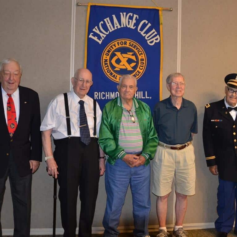 Fiver elderly men, one in a military uniform, posing in front of the Exchange Club banner