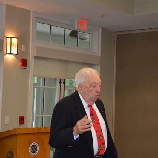 An elderly man walking away from a podium sporting a blue jacket and red patterned tie