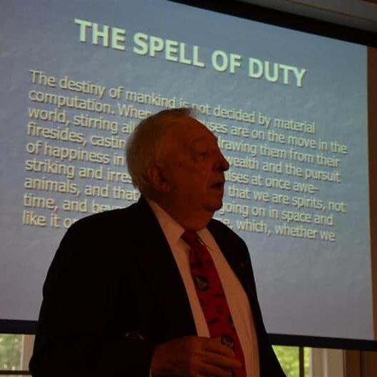 Elderly veteran standing in front of a PowerPoint screen entitled The Spell of Duty