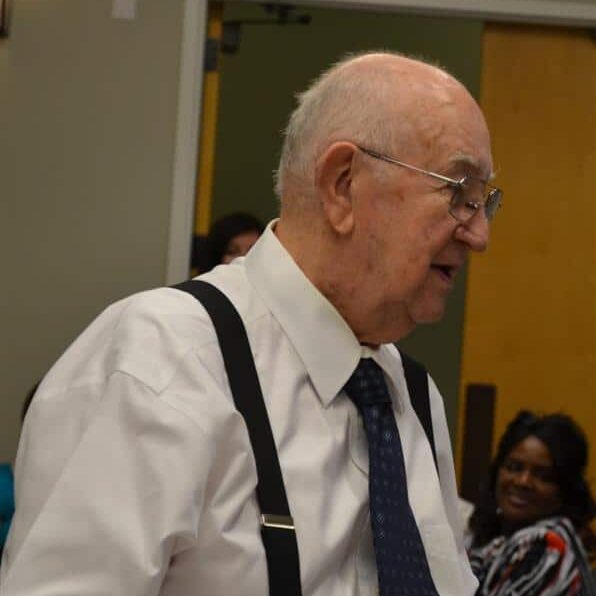 An elderly man wearing a white dress shirt, black suspenders, and a blue tie making is way from the podium back to his seat