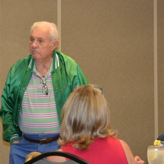 An elderly man in a green windbreaker talking to w women sitting at a banquet table