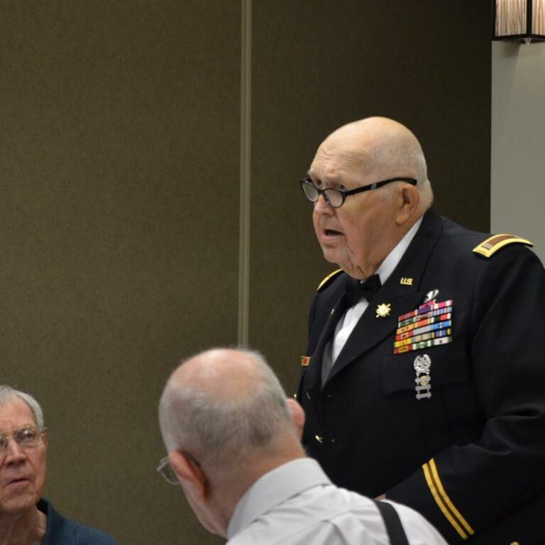 An older man in a military uniform decorated with badges and medals standing while talking to two fellow veterans